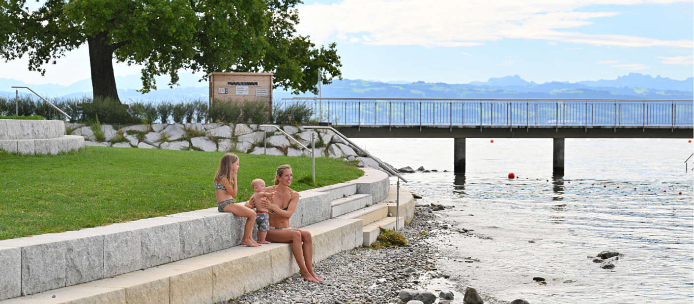 Mutter sitzt mit Mädchen und Baby auf Steinstufen am Bodenseeufer.